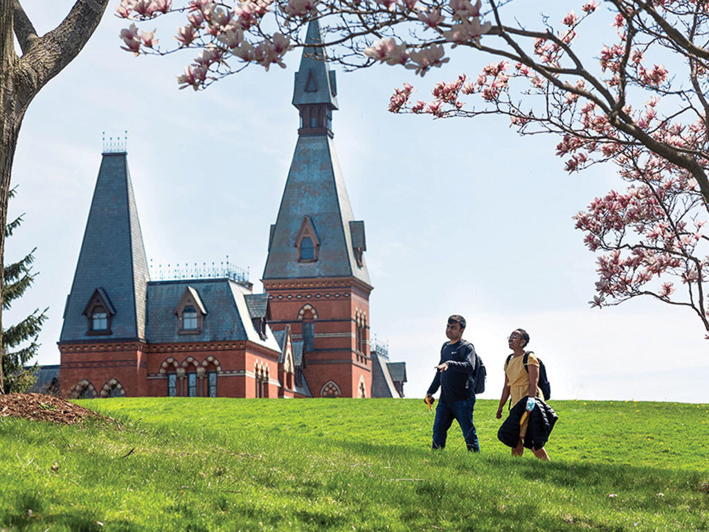 Two people walk across the Cornell campus with Sage Hall in the background.