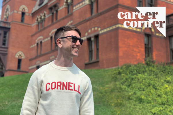 a young man wearing a Cornell sweatshirt and sunglasses standing in front of Sage Hall and smiling.