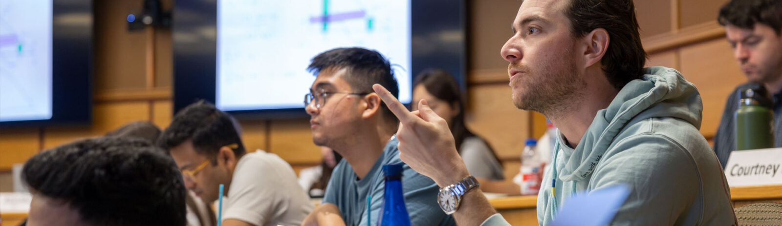 A student speaks and gestures with his pointer finger in a full tiered classroom.