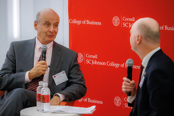 One man speaking into a microphone and looking at another man seated adjacent to him at a small table.